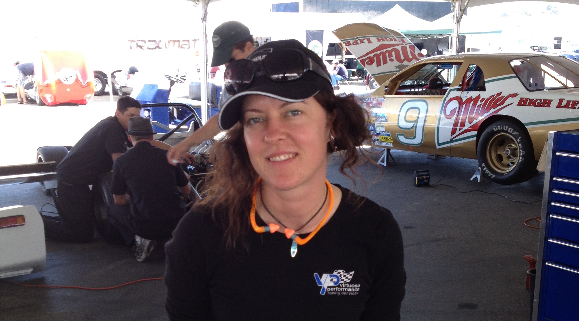 woman sitting on a stack of race wheels, wearing ear protection and safety glasses while in the background you can see a gold colored Nascar car jacked up with the hood open