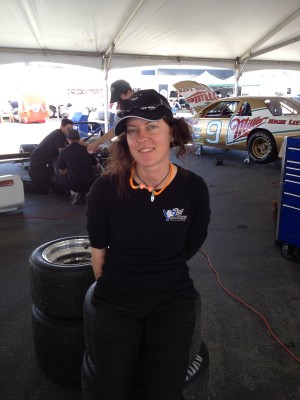 woman sitting on a stack of race wheels, wearing ear protection and safety glasses while in the background you can see a gold colored Nascar car jacked up with the hood open