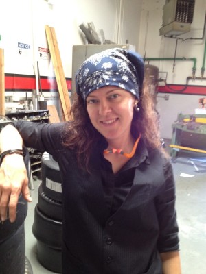 woman wearing a blue bandanna, dark long sleeved button down shirt under a pinstripe vest and ear protection hanging around her neck standing in the middle of a workshop