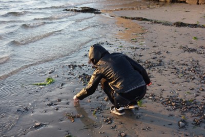 girl in leather jacket, on beach, facing away from camera, picking something off the sand near the water's edge