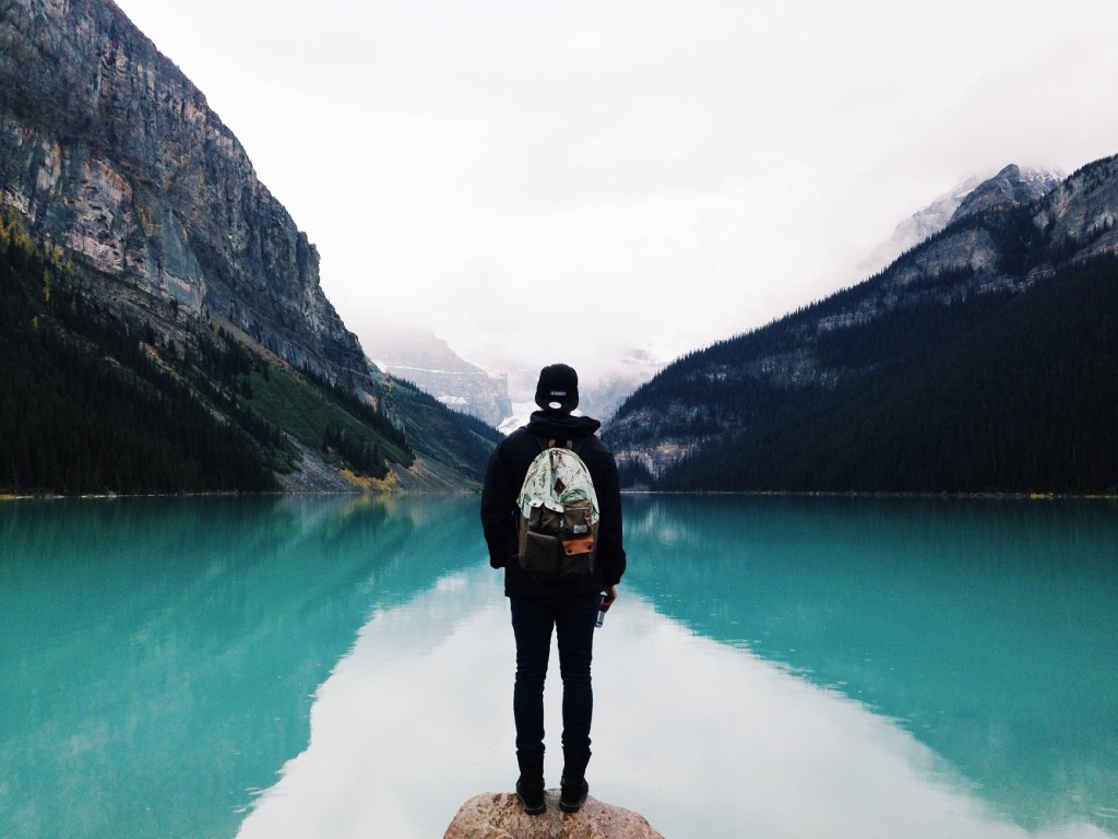 person facing away from camera, standing at the edge of a turquoise lake with a mountain rising out of the lake on the left and another on the right