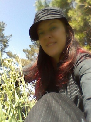 smiling woman with long red hair, wearing a black cap, black pinstripe pants and a black long sleeve button down shirt, sitting in tall grass with pine trees behind her.