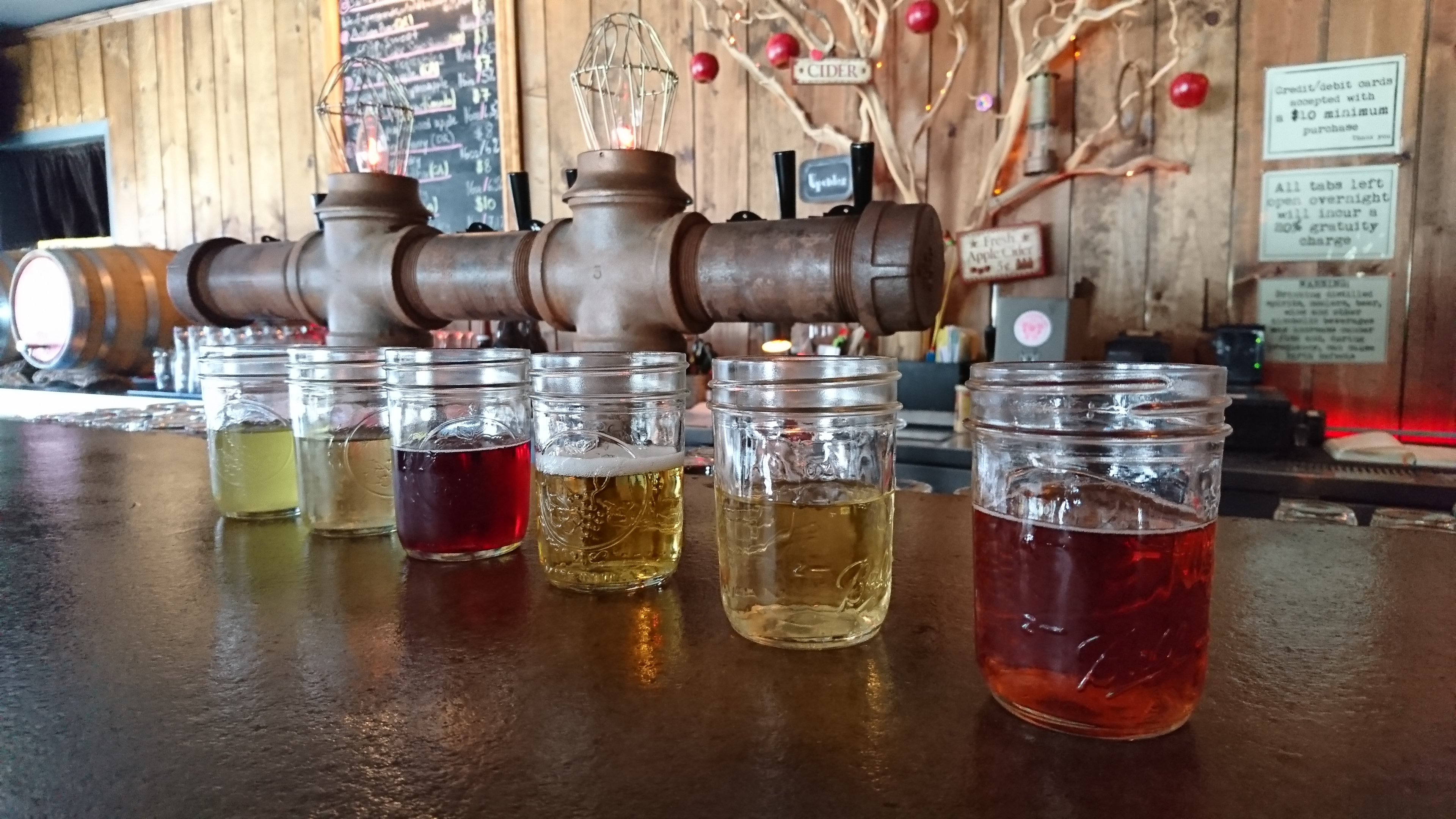 half full jars of red and yellow cider lined up on a copper colored bar with copper colored pipes behind the jars and bare branches pinned to the wall with apples hanging from the branches.