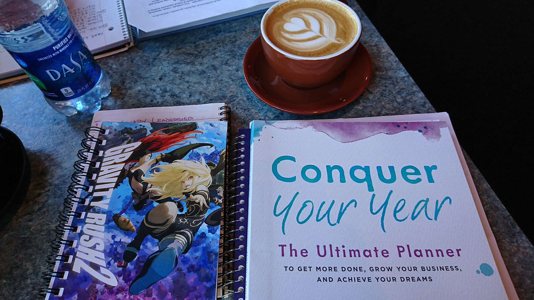 a cappuccino with a heart drawn in the milk next to books on a table. The books include notebooks and a yearly planner